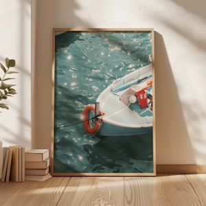 Op de afbeelding: Een ingelijste foto van een klein blauw bootje dat drijft op turquoise water. Het bootje heeft een reddingsboei aan de zijkant en een rode emmer erin. Het water glinstert in het zonlicht.