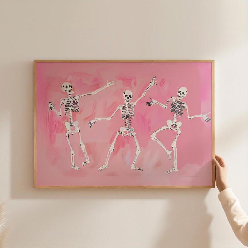 May include: A framed art print featuring three skeletons dancing on a pink background. The skeletons are painted in black and white, and the background is a soft, pastel pink.