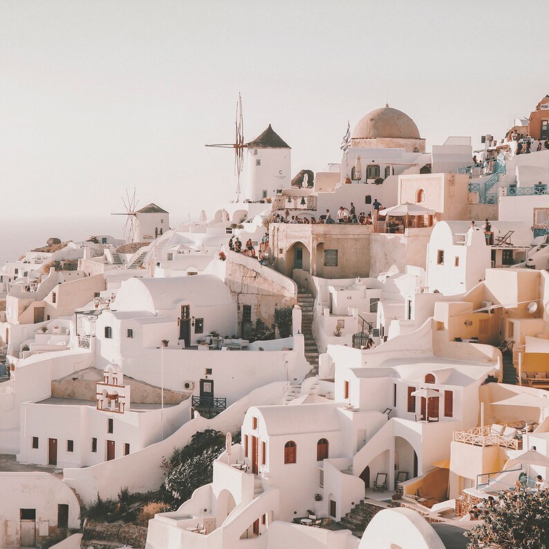 May include: A whitewashed village with a windmill on a hilltop. The buildings are clustered together and have a traditional Greek architecture. The sky is a light blue and the sun is shining.