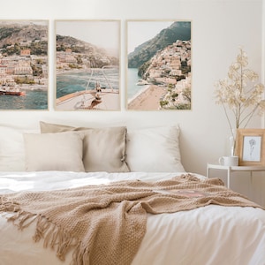 May include: Three framed photographs of a coastal town with colorful buildings on a hillside overlooking a blue ocean. The photos are hung on a white wall above a white bed with a beige blanket.