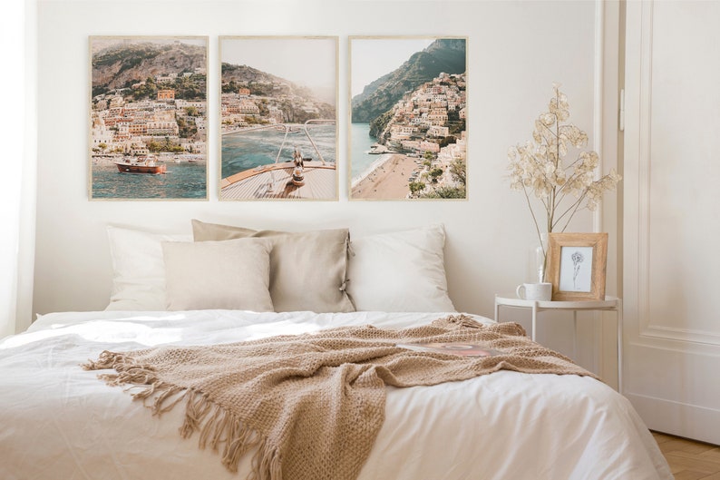 May include: Three framed photographs of a coastal town with colorful buildings on a hillside overlooking a blue ocean. The photos are hung on a white wall above a white bed with a beige blanket.