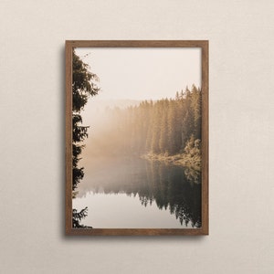 Op de afbeelding: Een ingelijste landschapsafdruk met een sereen meer dat een bos van hoge naaldbomen weerspiegelt. De afbeelding baadt in zacht, warm licht, met een nevelige sfeer. De lijst is van een middelbruine kleur.