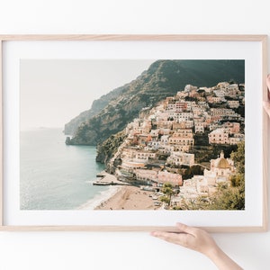 May include: A framed photograph of a colorful town built on a steep cliffside overlooking a blue ocean. The town is a mix of pastel pink, yellow, and white buildings with a few green trees. The ocean is a deep blue with a white sandy beach in the foreground.