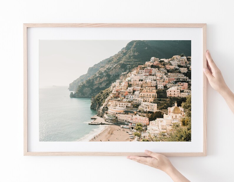 May include: A framed photograph of a colorful town built on a steep cliffside overlooking a blue ocean. The town is a mix of pastel pink, yellow, and white buildings with a few green trees. The ocean is a deep blue with a white sandy beach in the foreground.