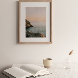 May include: A framed photograph of a rocky coastline with a sailboat in the distance. The sky is a soft pink and orange, and the water is a deep blue. The frame is made of light wood.