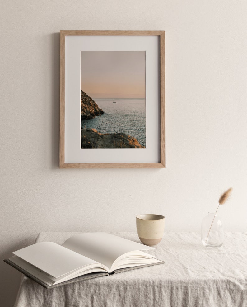 May include: A framed photograph of a rocky coastline with a sailboat in the distance. The sky is a soft pink and orange, and the water is a deep blue. The frame is made of light wood.