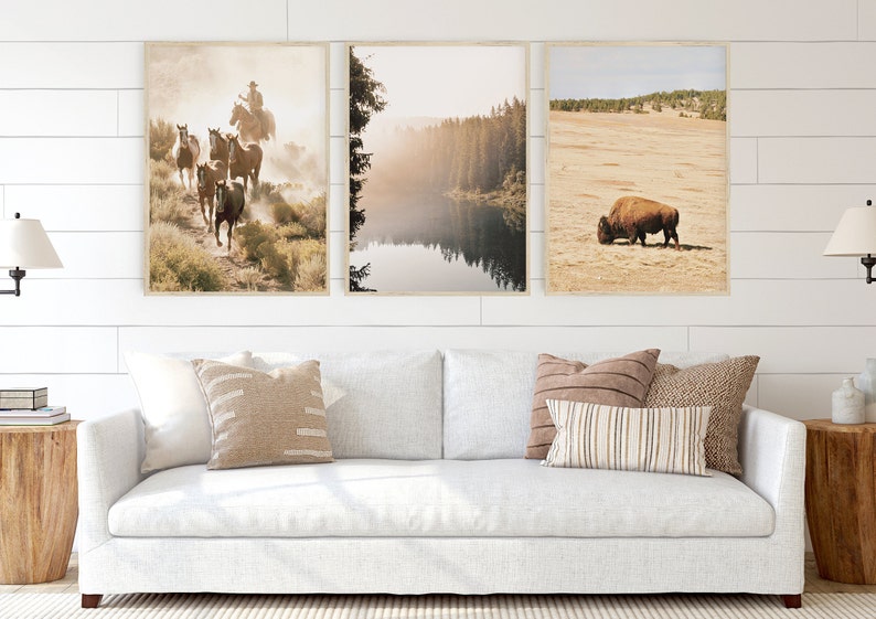 Op de afbeelding: Drie ingelijste landschapsprints. De eerste toont een cowboy die paarden berijdt op een stoffig pad. De tweede toont een meer omgeven door bomen. De derde toont een bizon die in een veld graast. De prints worden boven een witte bank weergegeven.