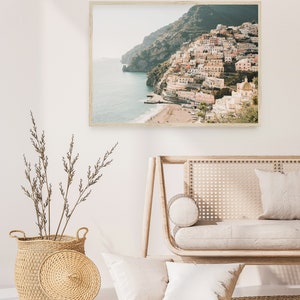 May include: A framed photograph of a coastal town with colorful buildings built into a hillside overlooking the ocean. The photo is displayed on a white wall above a light wood couch with white cushions.