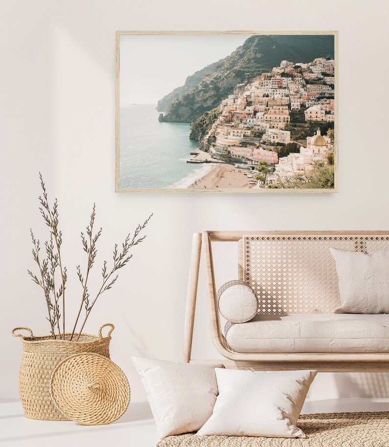 May include: A framed photograph of a coastal town with colorful buildings built into a hillside overlooking the ocean. The photo is displayed on a white wall above a light wood couch with white cushions.