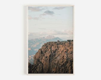 May include: A photograph of a rocky cliff overlooking a mountain range. The sky is a light blue with white clouds. The mountain range is in the distance and is covered in snow. The cliff is brown and rocky.