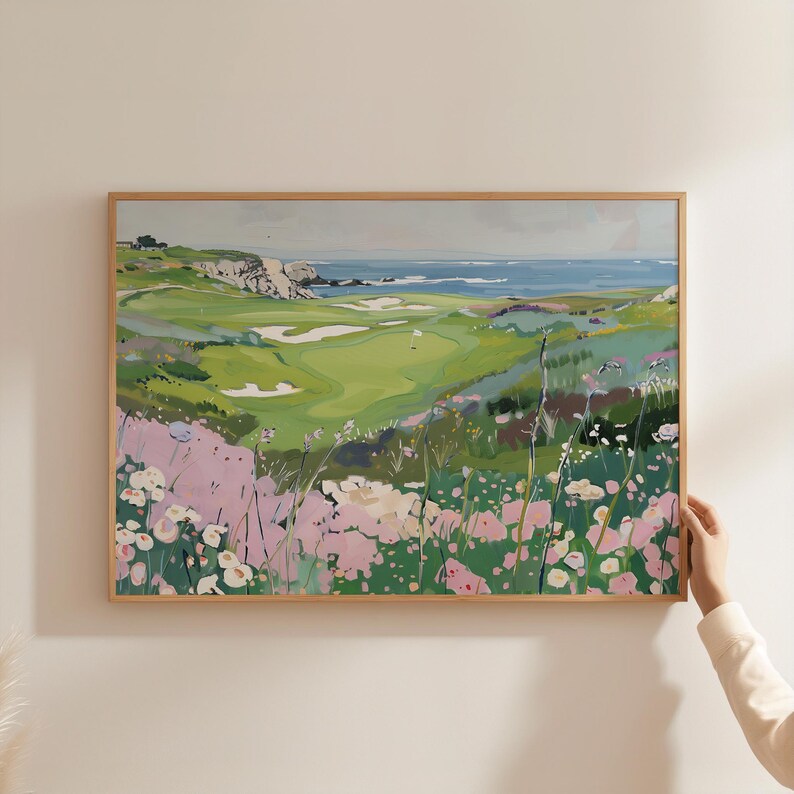 May include: A framed landscape painting of a golf course with a view of the ocean. The artwork features a green golf course, white sand traps, and a blue ocean. Pink and white flowers are in the foreground.