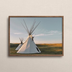 May include: Two white teepees stand in a grassy field against a blue sky with white clouds. The teepees are made of canvas and have wooden poles. The teepees are in a field of tall grass.