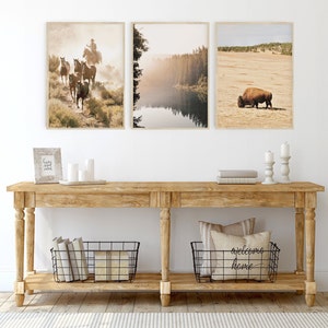 Op de afbeelding: Drie ingelijste landschapsprints met een cowboy te paard, een mistige meer sc&egrave;ne en een grazende bizon. Ze worden tentoongesteld boven een houten consoletafel met decoratieve items en opbergmanden.