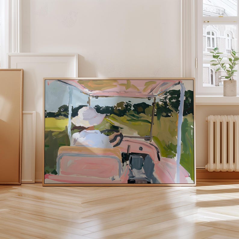 May include: Abstract painting of a golf cart in a landscape. The artwork features a pink golf cart with a person wearing a white hat. The background includes green trees and a yellow field. The painting is framed in a light wood frame.
