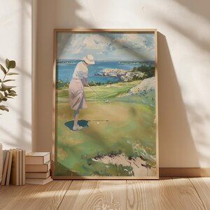 May include: A framed golf-themed art print. The artwork depicts a person in a white shirt and pink skirt playing golf on a green course with a blue ocean and sky in the background. The print is in a light brown wooden frame.
