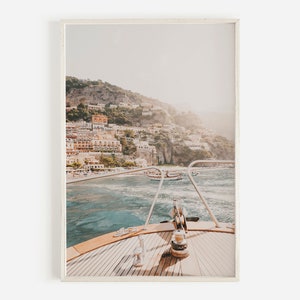 May include: A view from the bow of a boat, looking out at a coastal town built into a hillside. The water is a light blue and the sky is a pale blue. The town is a mix of white and yellow buildings.