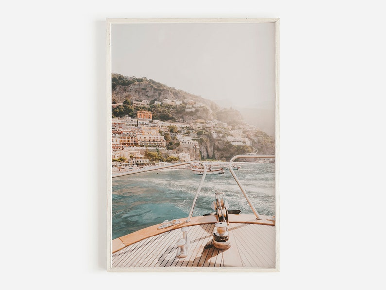 May include: A view from the bow of a boat, looking out at a coastal town built into a hillside. The water is a light blue and the sky is a pale blue. The town is a mix of white and yellow buildings.