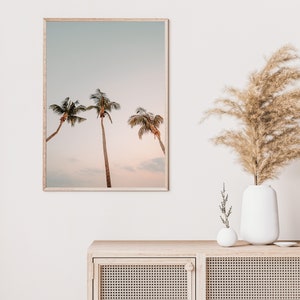May include: A framed print of three palm trees against a soft, pastel sky. The sky is a light blue with hints of pink and orange. The palm trees are silhouetted against the sky.