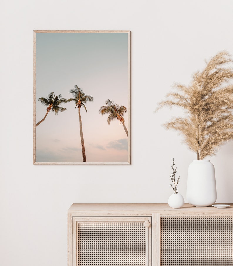 May include: A framed print of three palm trees against a soft, pastel sky. The sky is a light blue with hints of pink and orange. The palm trees are silhouetted against the sky.
