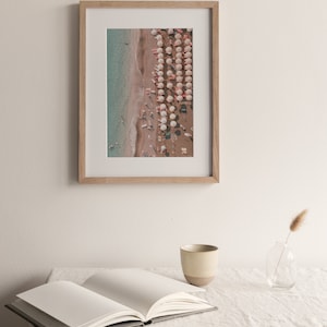 Peut inclure: Une photographie encadr&eacute;e d'une sc&egrave;ne de plage avec des parasols blancs et des gens sur le sable. L'eau est d'un bleu clair et le sable est d'un brun clair.