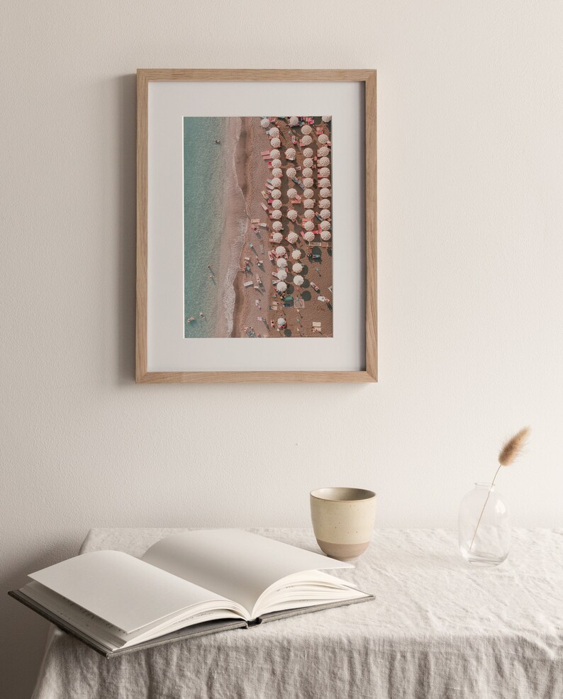 Peut inclure: Une photographie encadr&eacute;e d'une sc&egrave;ne de plage avec des parasols blancs et des gens sur le sable. L'eau est d'un bleu clair et le sable est d'un brun clair.
