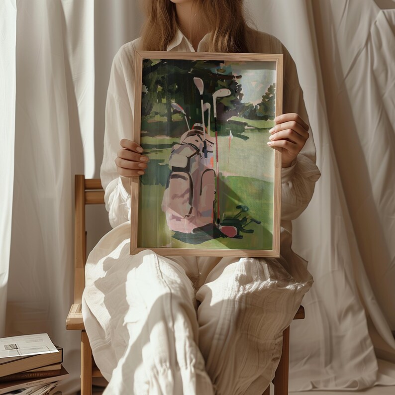 May include: A framed painting of a golf course scene. The artwork shows a pink golf bag with clubs on a green fairway, with trees in the background. The painting is held by a person wearing a white shirt and trousers.