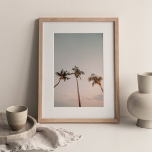 May include: A framed print of three palm trees against a light blue sky with a pink sunset in the background. The frame is made of light wood.