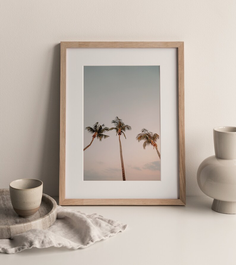 May include: A framed print of three palm trees against a light blue sky with a pink sunset in the background. The frame is made of light wood.