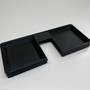 May include: A black, rectangular tray with a unique, angular design. The tray features two main compartments and a smaller, connecting section. The surface has a textured pattern, and the overall shape is reminiscent of a stylized letter 'L'.