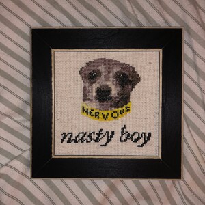 Puede incluir: Un punto de cruz enmarcado de un perro con un collar amarillo que dice "NERVOUS" y el texto "nasty boy" debajo del perro.