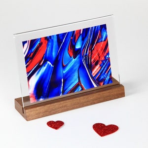 May include: A framed abstract art print with swirling blue and red paint. The print is displayed on a wooden stand with two red glitter hearts in the foreground.