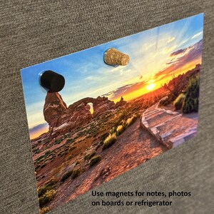 May include: A sunset landscape photograph with a rock formation in the foreground. The photo is attached to a gray bulletin board with two magnets, one black and one brown.
