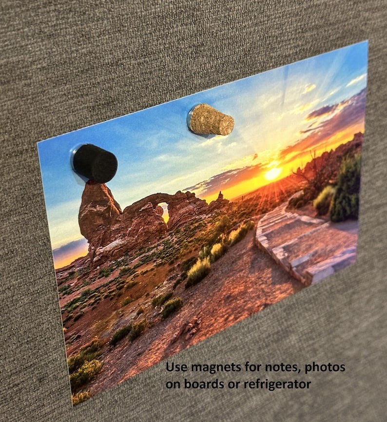 May include: A sunset landscape photograph with a rock formation in the foreground. The photo is attached to a gray bulletin board with two magnets, one black and one brown.
