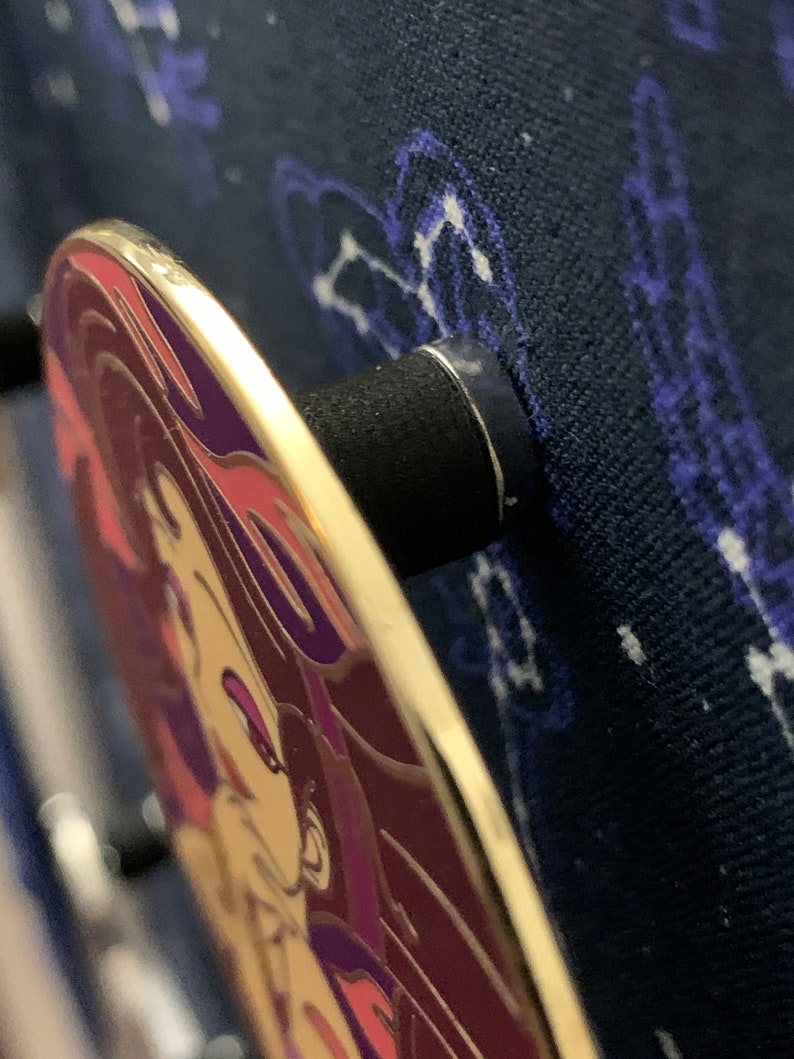 May include: A gold-colored enamel pin with a purple and pink design of a woman's face. The pin is attached to a black backing.