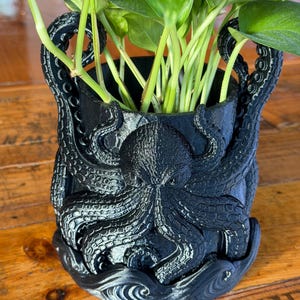 May include: Black octopus-themed planter pot holding a green leafy plant. The planter features a detailed octopus design with tentacles wrapping around the pot. The pot sits on a wooden surface.