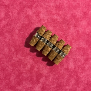 May include: A group of eight cork bottle stoppers with a silver metal band around each cork. The stoppers are arranged in a staggered pattern.