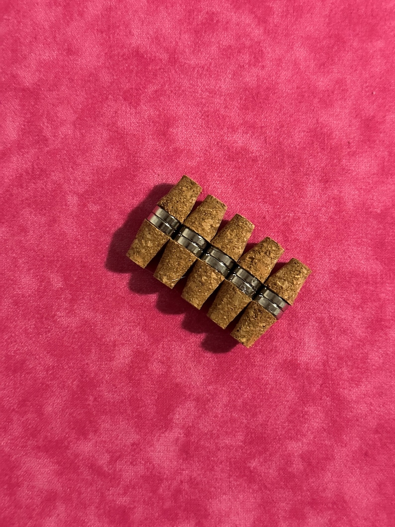 May include: A group of eight cork bottle stoppers with a silver metal band around each cork. The stoppers are arranged in a staggered pattern.