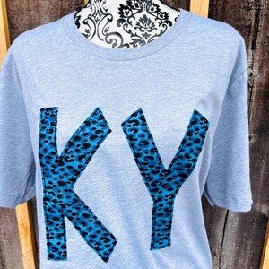 May include: A light blue t-shirt featuring the letters "KY" in a large, bold font. The letters are made from a blue and black leopard print fabric.