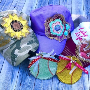 May include: Three embroidered baseball caps with different designs. The first cap is camouflage with a yellow sunflower and the text "Hello Sunshine". The second cap is purple with a colourful flower. The third cap is white with a red anchor and the text "Lake Life". Two glitter baseball keychains are also shown.