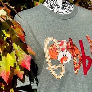 Peut inclure: Un t-shirt vert avec une applique de Thanksgiving repr&eacute;sentant une dinde avec le mot "Gobble" en motif &eacute;cossais.
