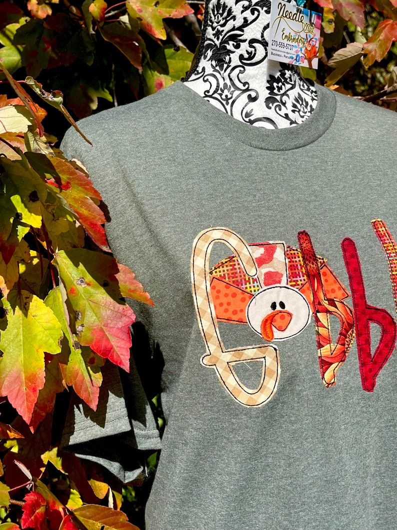 Peut inclure: Un t-shirt vert avec une applique de Thanksgiving repr&eacute;sentant une dinde avec le mot "Gobble" en motif &eacute;cossais.