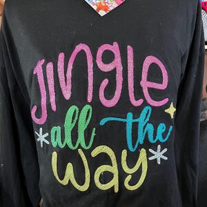 May include: Black long-sleeve shirt with the phrase "Jingle all the way" embroidered in colorful, glittery thread. The text includes pink, green, blue, and yellow hues, with star and snowflake accents. A tag is attached to the shirt.