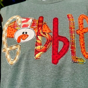Peut inclure: Un t-shirt gris avec le mot "Gobble" &eacute;crit en lettres de tissu color&eacute;es. La lettre "G" est une dinde avec un bec et une barbichette rouges.