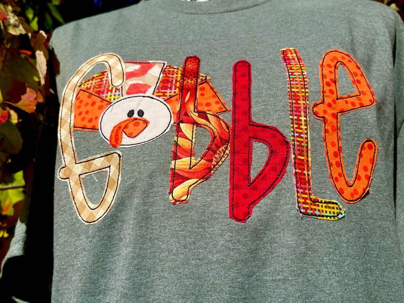 Peut inclure: Un t-shirt gris avec le mot "Gobble" &eacute;crit en lettres de tissu color&eacute;es. La lettre "G" est une dinde avec un bec et une barbichette rouges.