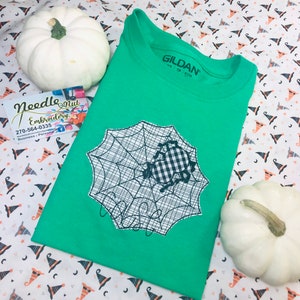 May include: A green t-shirt featuring a black and white spider web design with a gingham spider applique. The shirt is folded and placed on a patterned fabric with white pumpkins. The brand name "GILDAN" is visible on the shirt's collar.