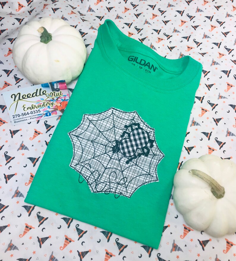 May include: A green t-shirt featuring a black and white spider web design with a gingham spider applique. The shirt is folded and placed on a patterned fabric with white pumpkins. The brand name "GILDAN" is visible on the shirt's collar.