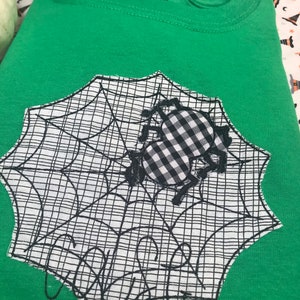 May include: Green t-shirt with a black and white spider web design and a gingham spider applique. The word "Creepy" is embroidered in black on the web. The shirt is folded.