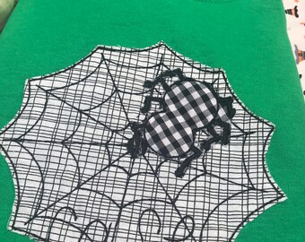 Puede incluir: Camiseta verde con un diseño de telaraña en blanco y negro y una aplicación de araña a cuadros. La palabra "Creepy" está bordada en negro en la telaraña. La camiseta está doblada.