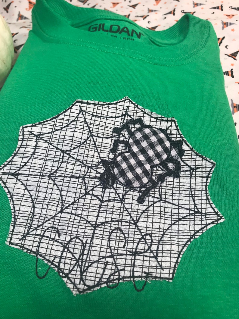 May include: Green t-shirt with a black and white spider web design and a gingham spider applique. The word "Creepy" is embroidered in black on the web. The shirt is folded.