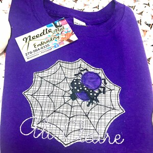 May include: A purple sweatshirt featuring a spider web design with a spider and the name "Claire" embroidered in white. The sweatshirt has a tag from "Needle Nut Embroidery". The background is a Halloween-themed fabric.